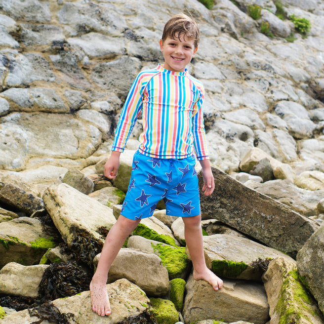 A boy smiling on rocks wearing UV Protective Swim Shorts Blue Starfish and a striped top, looking at the camera.